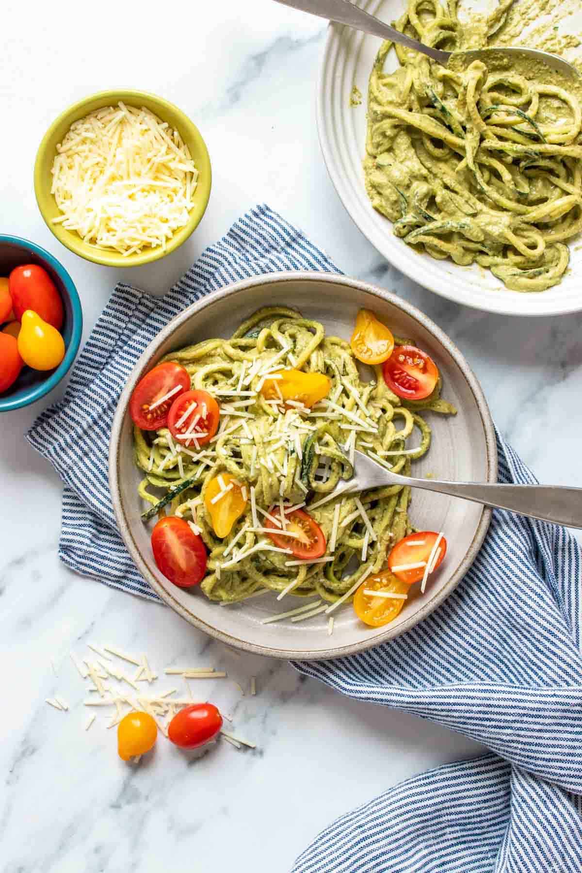 A grey plate on a blue striped towel with zucchini noodles mixed with pesto and tomatoes