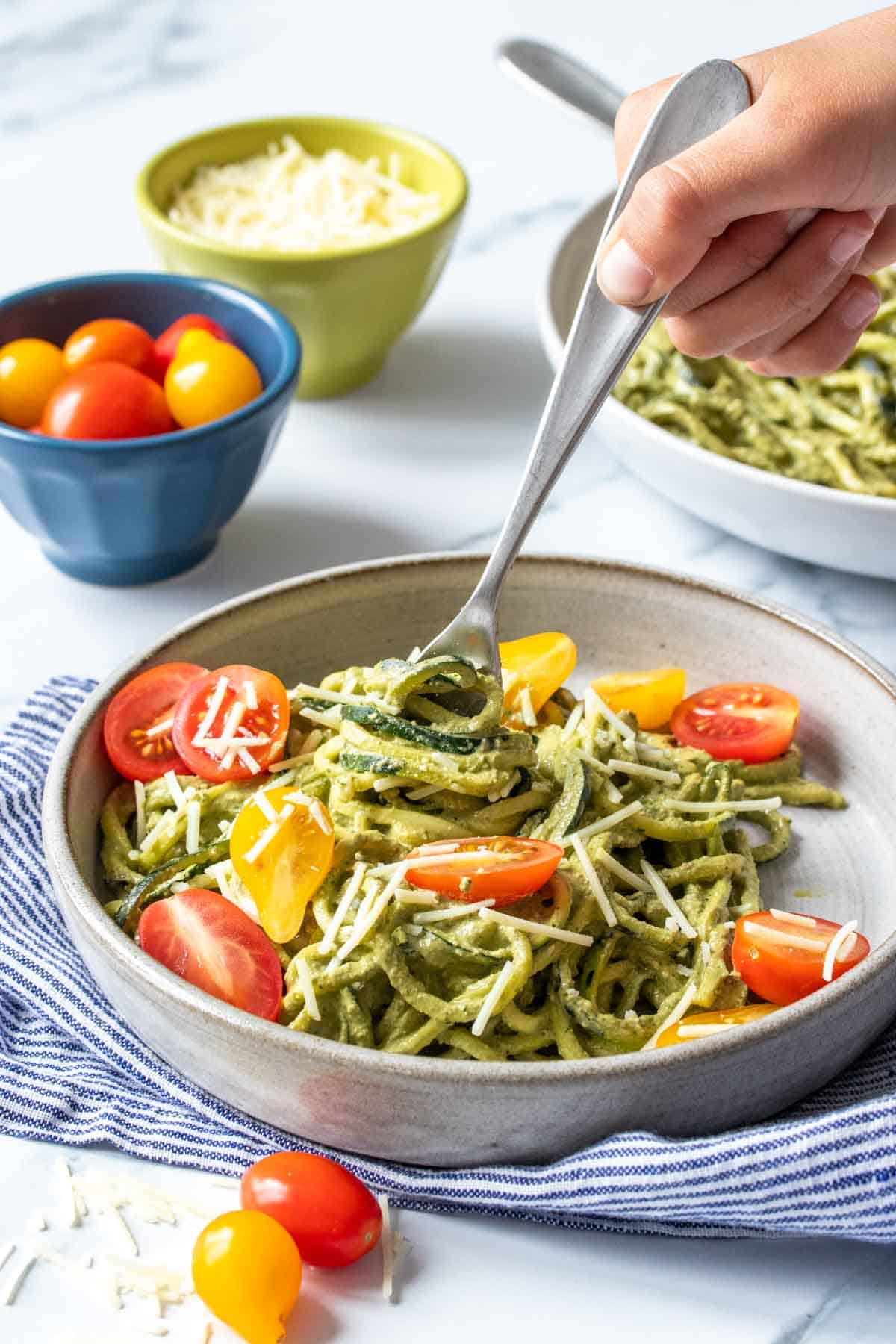 A hand holding a fork that is twisting pesto covered zucchini noodles onto it from a grey plate with tomatoes too