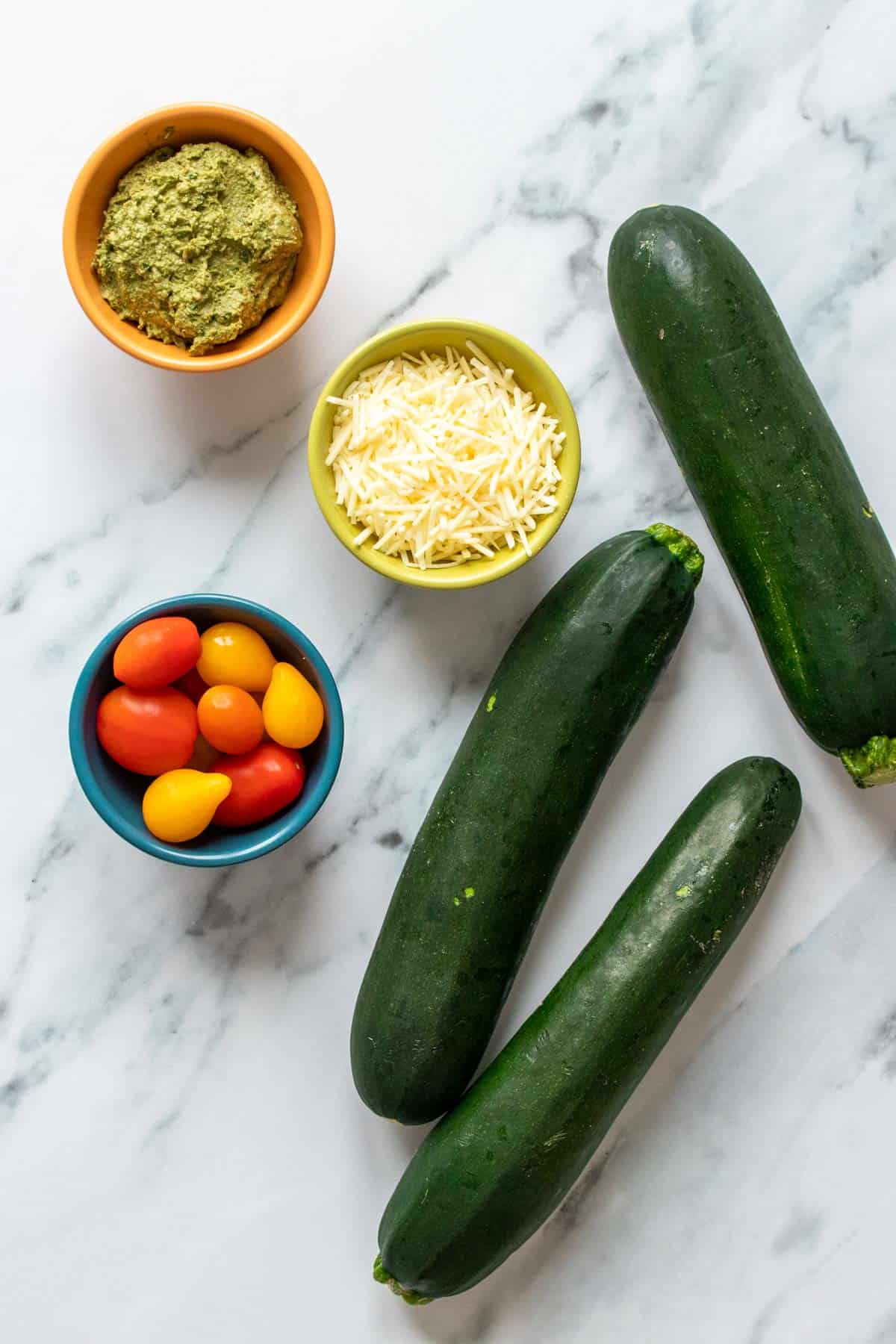 Top view of zucchini and bowls of parmesan, pesto and tomatoes on a marble surface