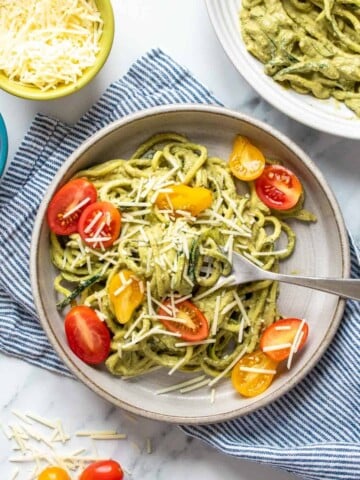 Pesto covered zucchini noodles topped with sliced tomatoes and parmesan on a grey plate with a fork on it