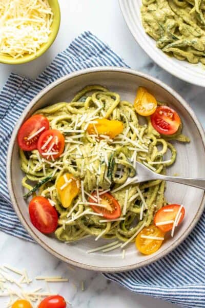 Pesto covered zucchini noodles topped with sliced tomatoes and parmesan on a grey plate with a fork on it