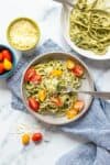 A grey plate with a fork on it and pesto zucchini noodles topped with sliced tomatoes and parmesan