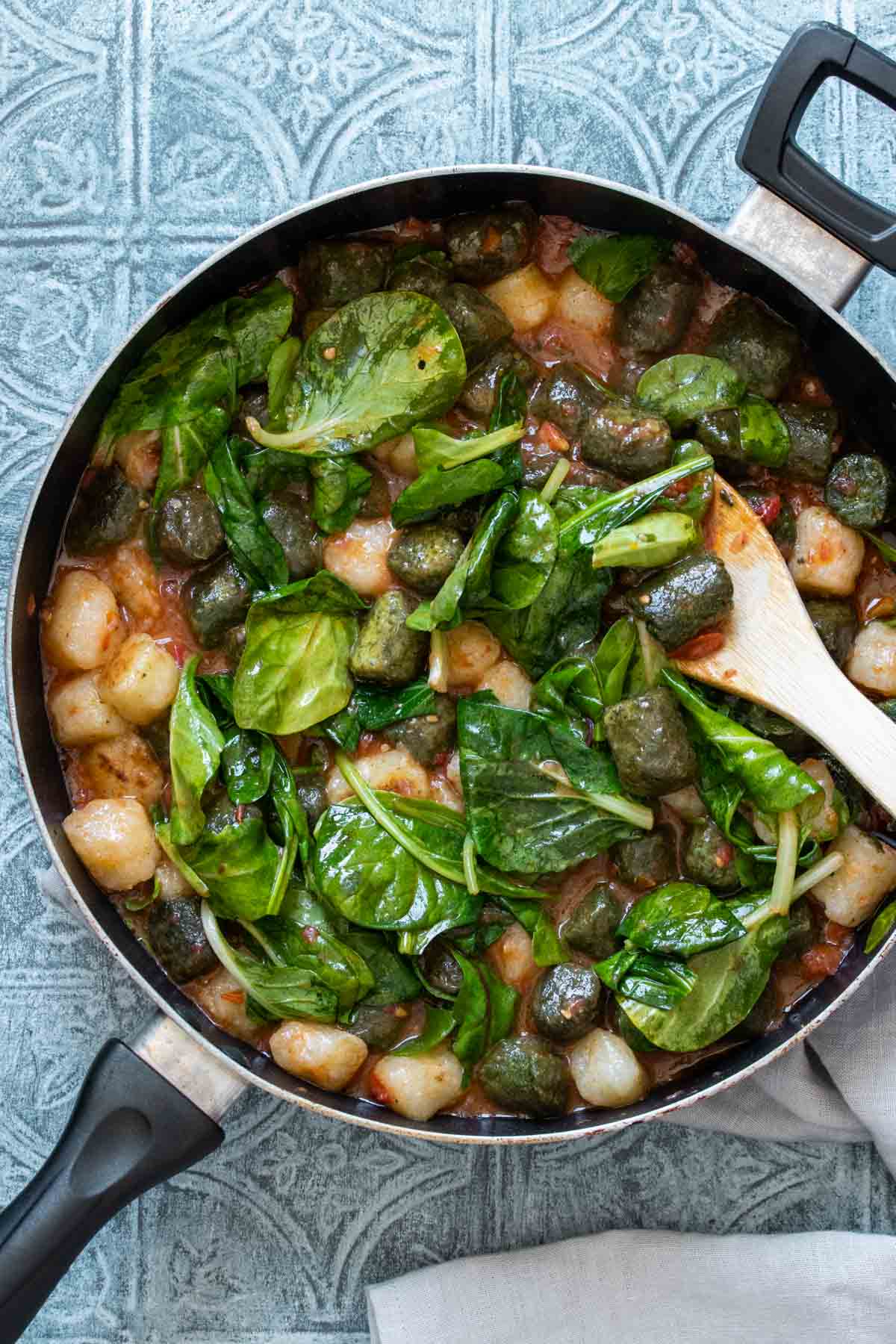 Fresh spinach on top of a mixture of gnocchi and red sauce in a pan being mixed by a wooden spoon