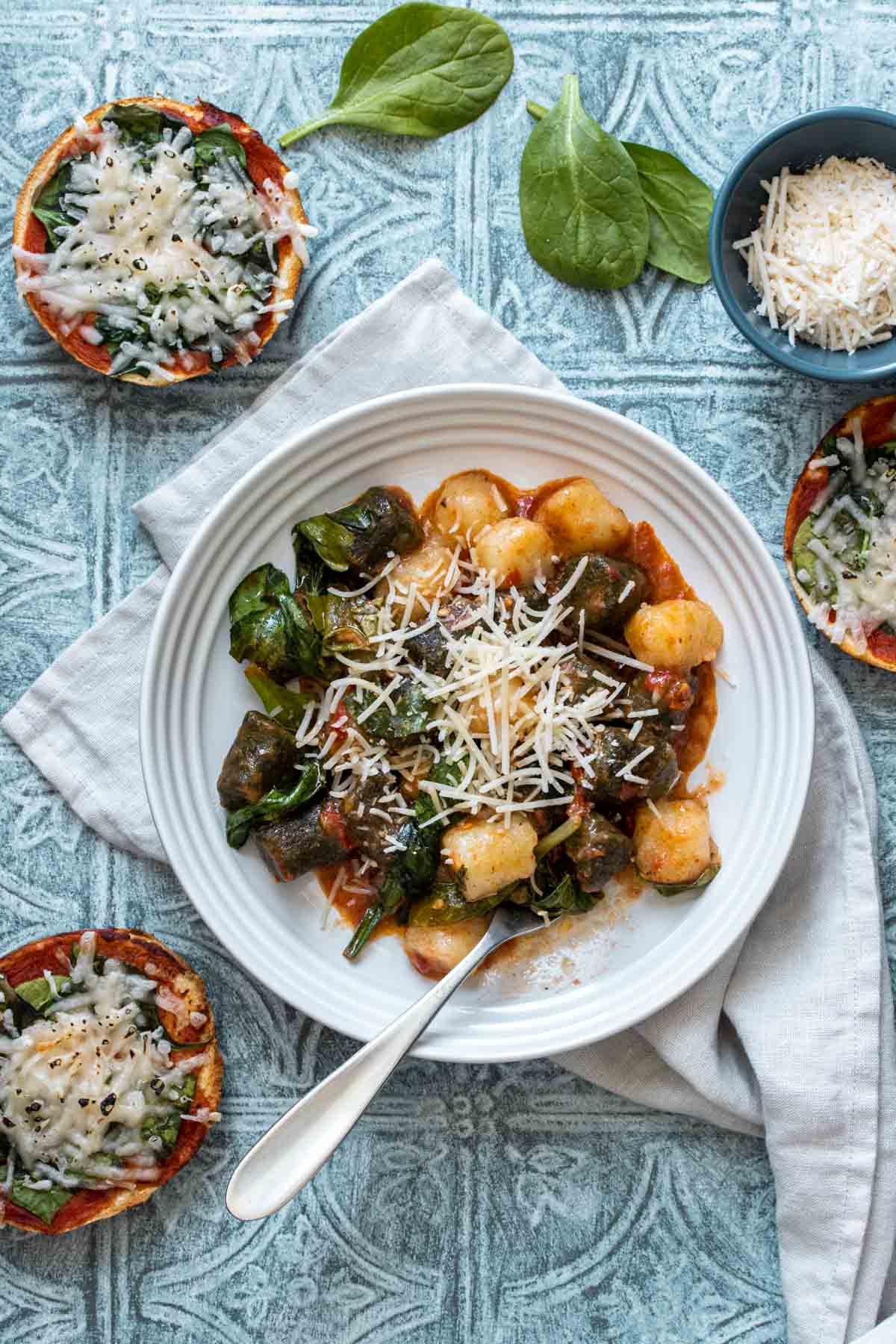 A white bowl with gnocchi, red sauce, spinach and parmesan on a blue surface with pizza bagels around it