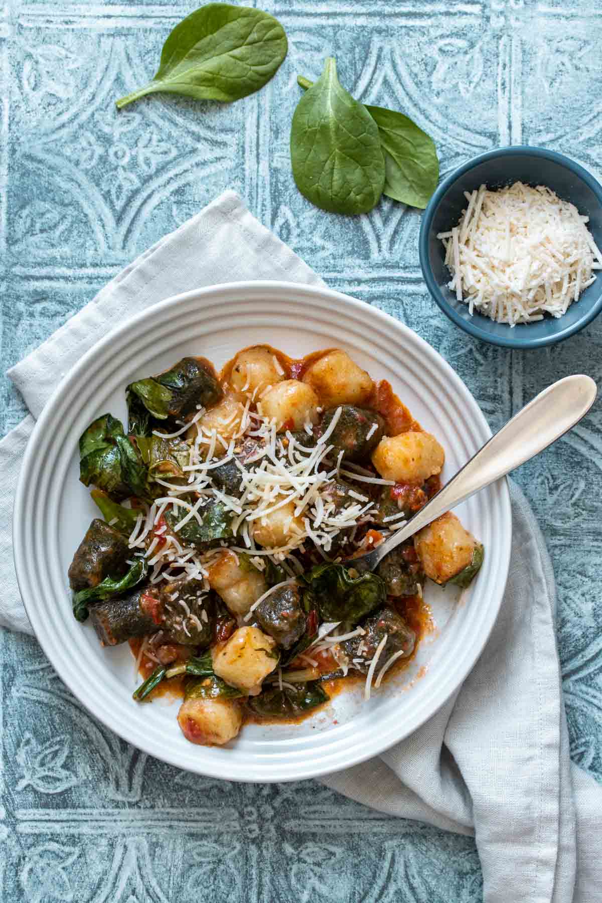 A fork in a white bowl with green and white gnocchi mixed with red sauce and spinach and topped with Parmesan.