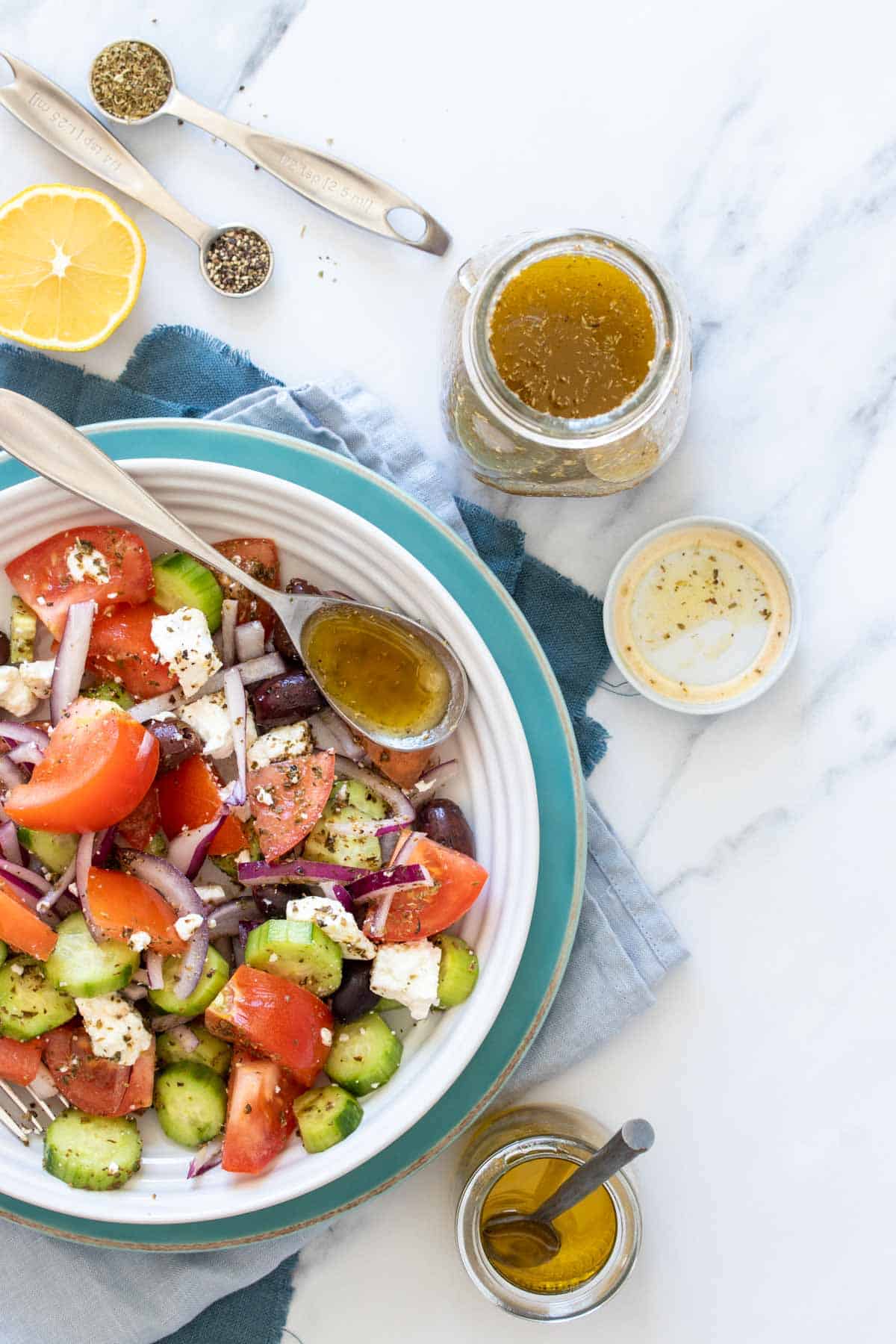 Top view of a Greek salad next to a jar of dressing with a spoon with dressing in it sitting on the plate