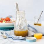 A glass salad dressing jar with a Greek dressing inside sitting on a blue towel next to a lemon and the jar lid.