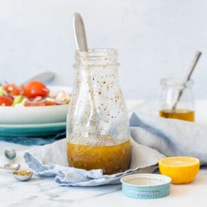 A glass salad dressing jar with a Greek dressing inside sitting on a blue towel next to a lemon and the jar lid.