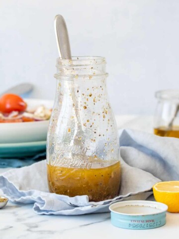 A glass salad dressing jar with a Greek dressing inside sitting on a blue towel next to a lemon and the jar lid.