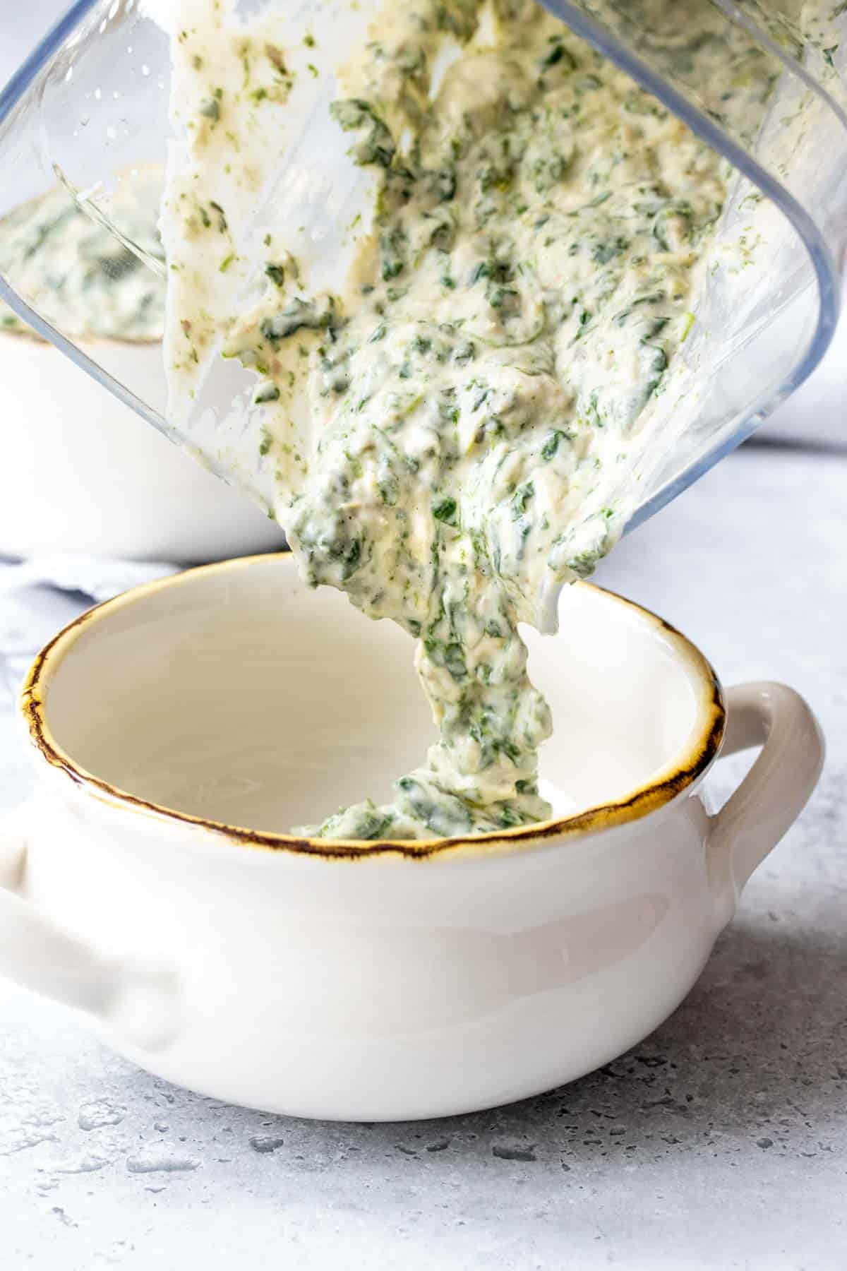 A blender pouring a spinach artichoke creamy dip mixture into a white ceramic bowl with handles
