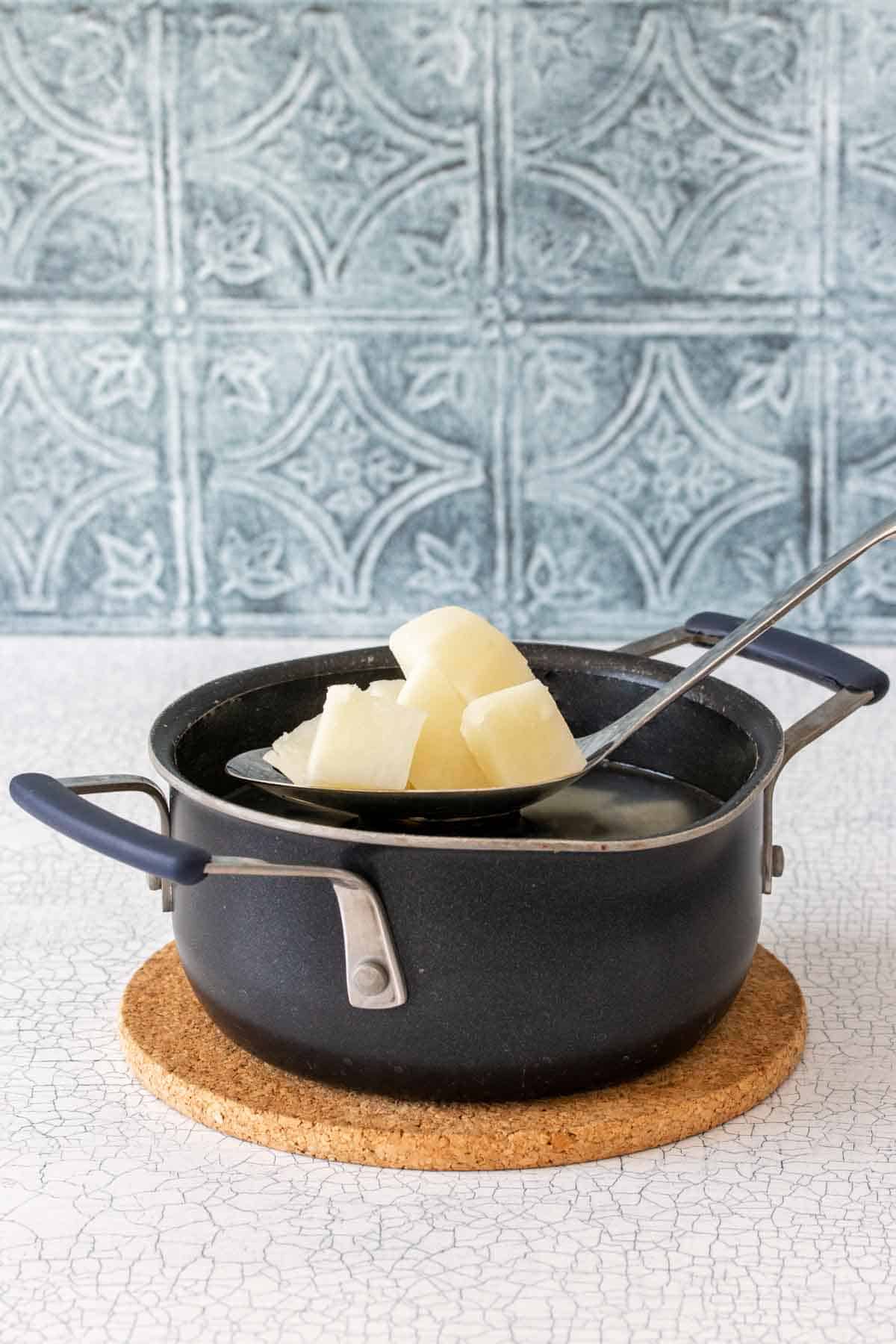 A slotted spoon with potato pieces coming out of a black pot with handles sitting on a cork trivet