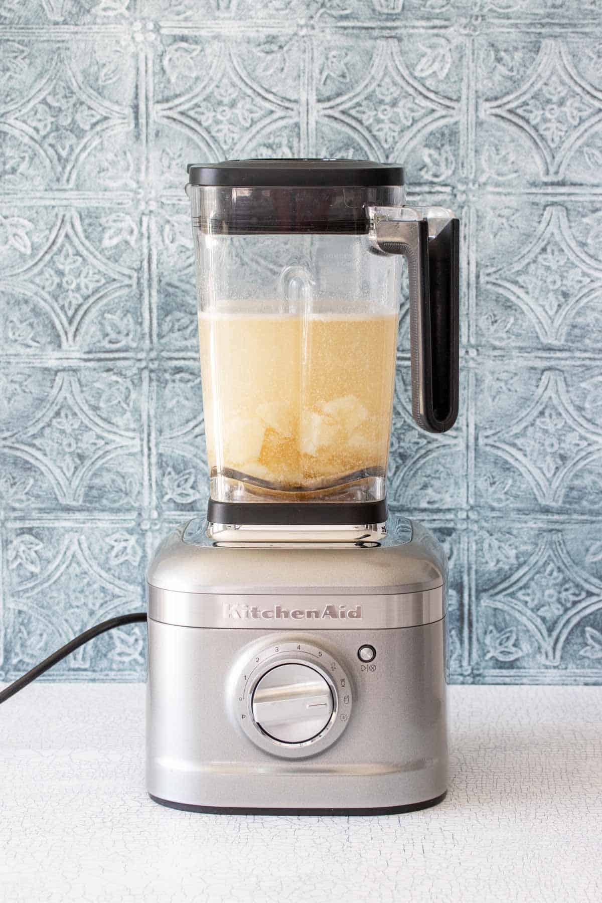 A white counter with a blue tile backsplash and a silver blender on it filled with potato pieces and a murky liquid