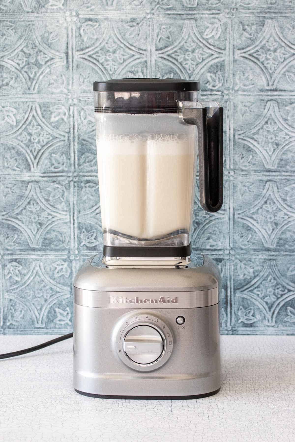 A silver blender filled with a milky liquid on a white counter with a blue tile background