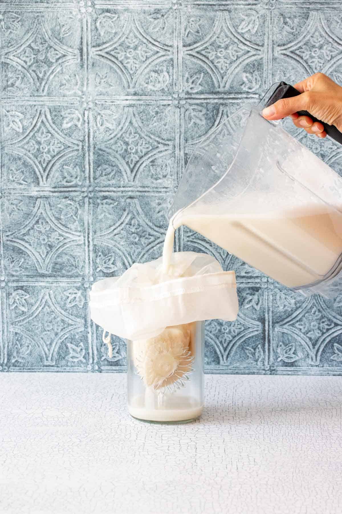A hand pouring milk from a blender into a strainer bag in a glass jar