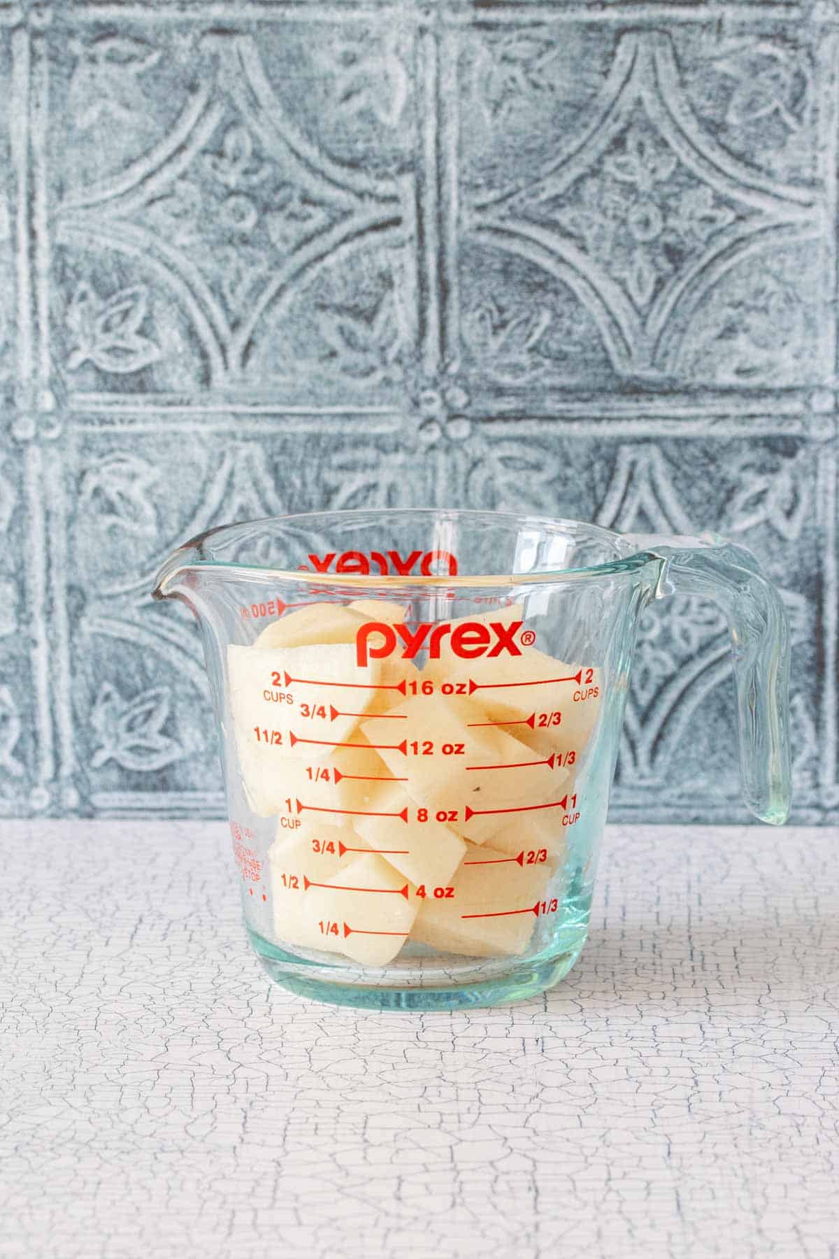 A glass pyrex measuring cup with peeled cut potatoes in it on a white counter in front of blue tile