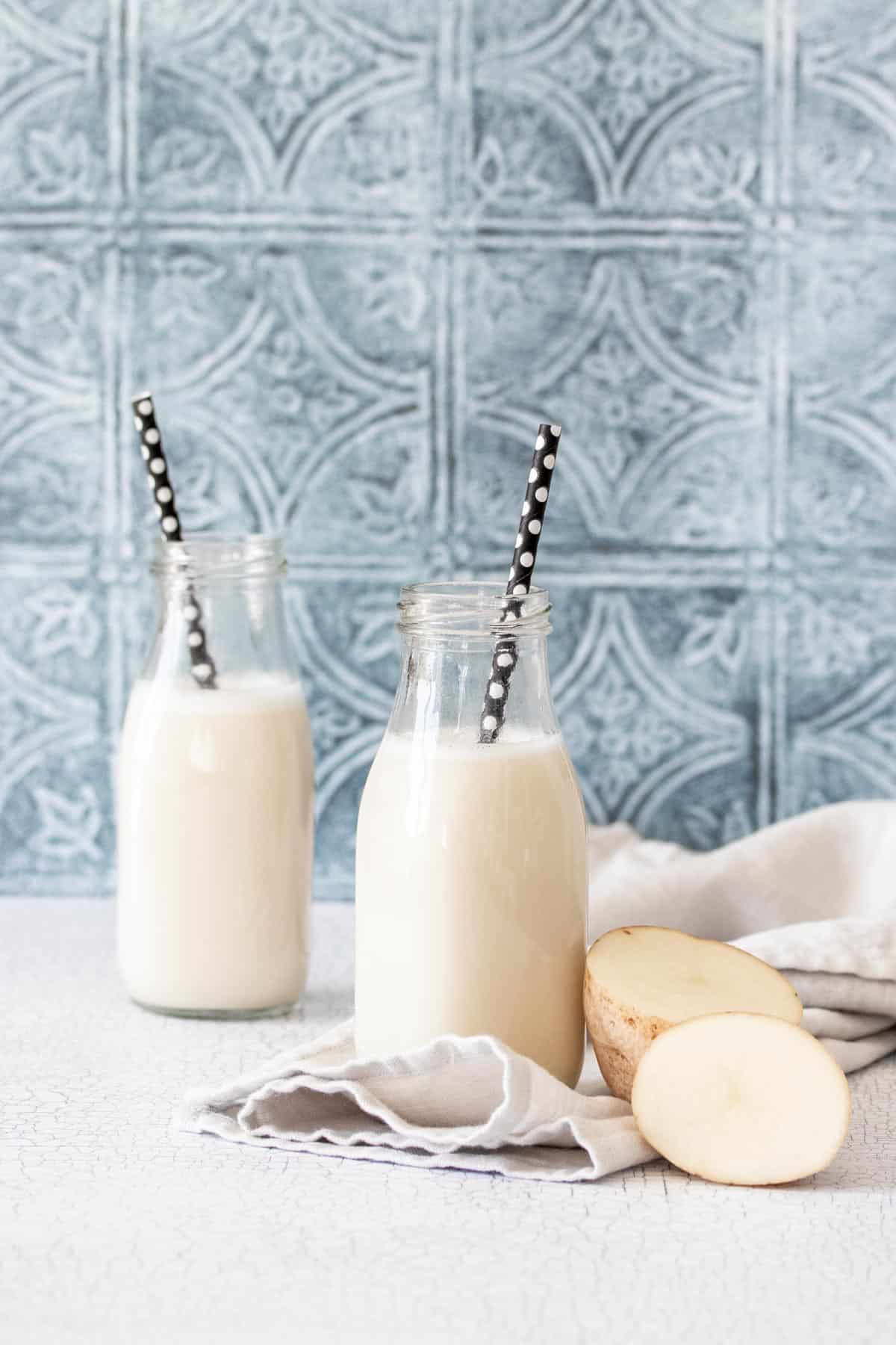 Two glass milk jars filled with milk and black straws with a cut potato next to the one in front on a kitchen counter