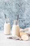 Two glass milk jars with black and white straws and a cut potato next to them on a counter with a tile background