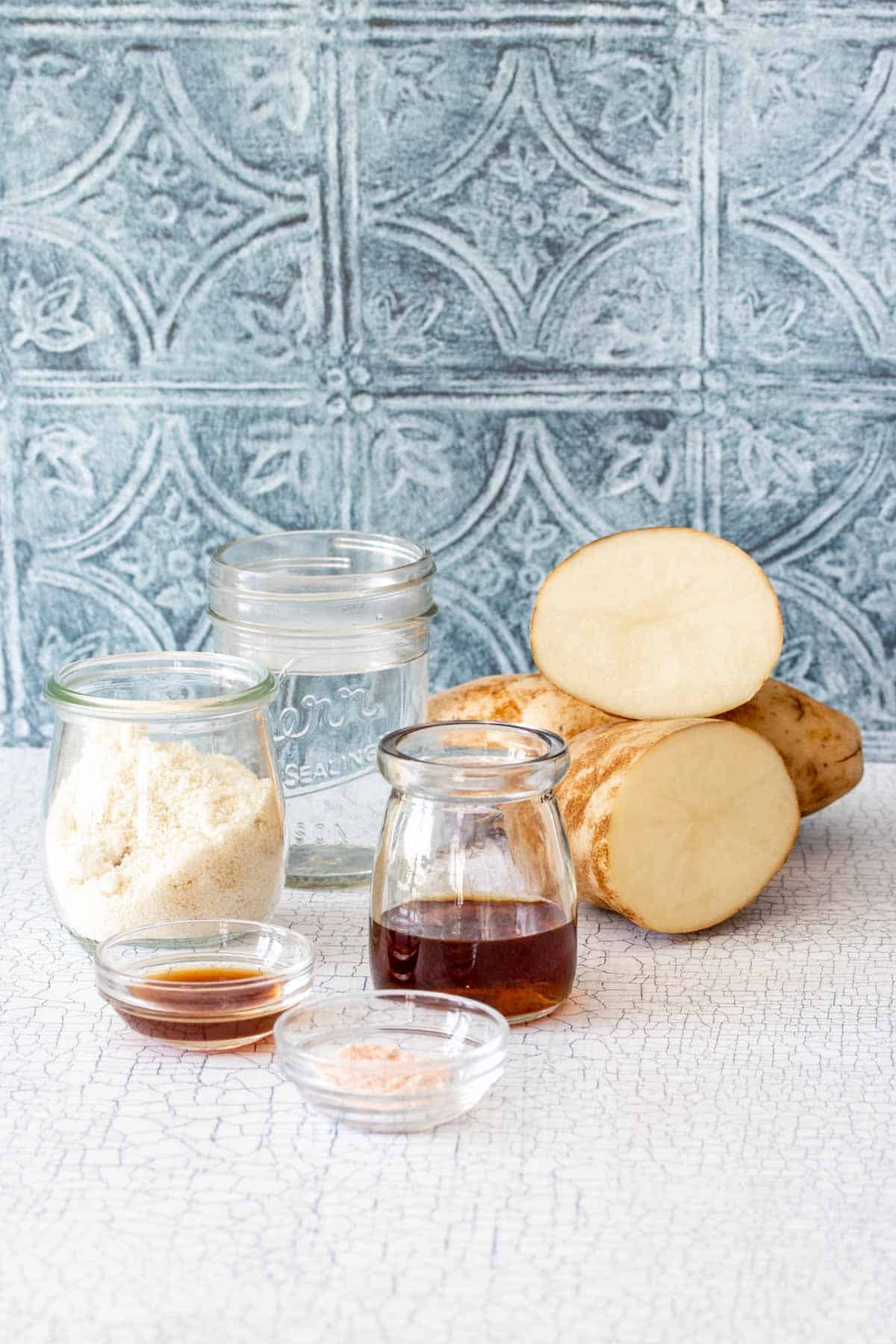 Potatoes and glass containers of flour, water, maple syrup, salt and vanilla on a white surface with a blue tile background