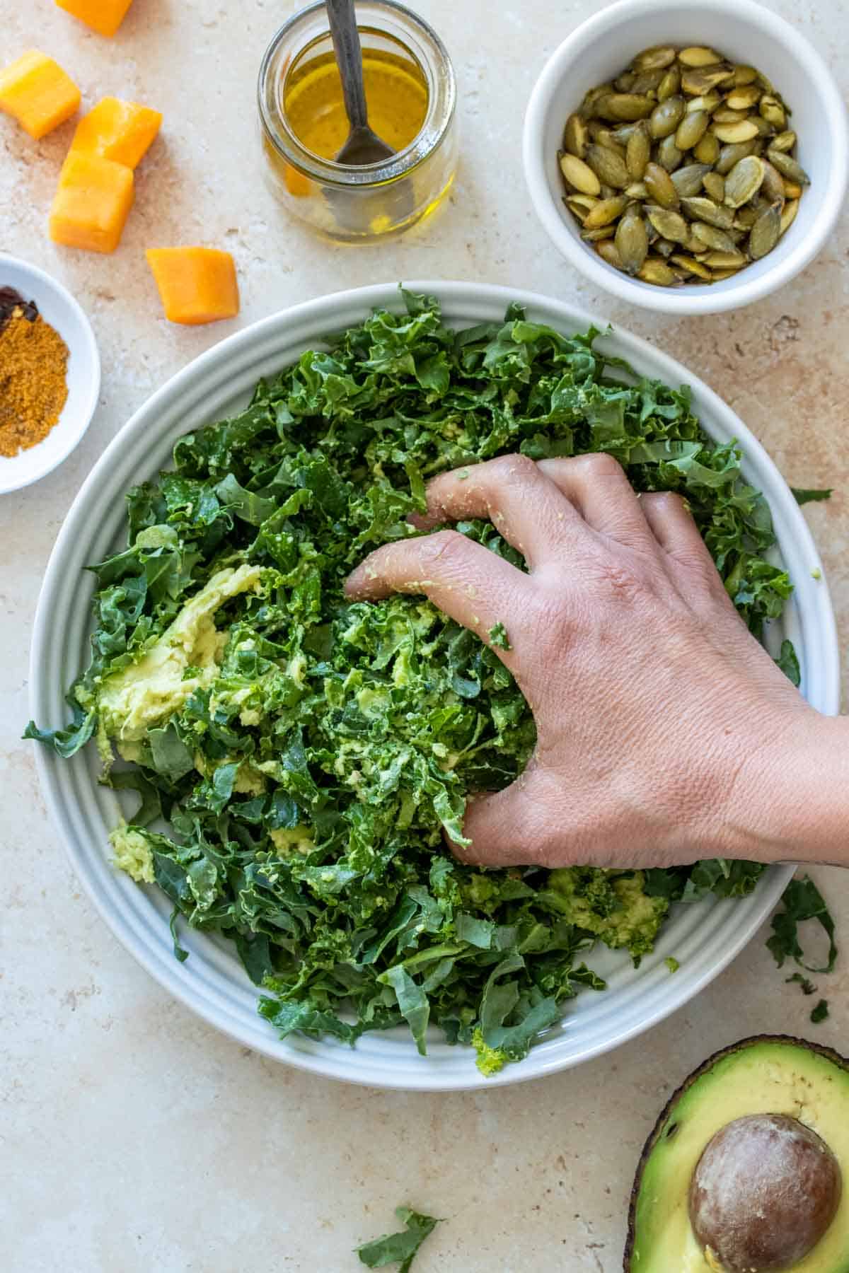A hand massaging avocado into a shredded kale salad in a white bowl