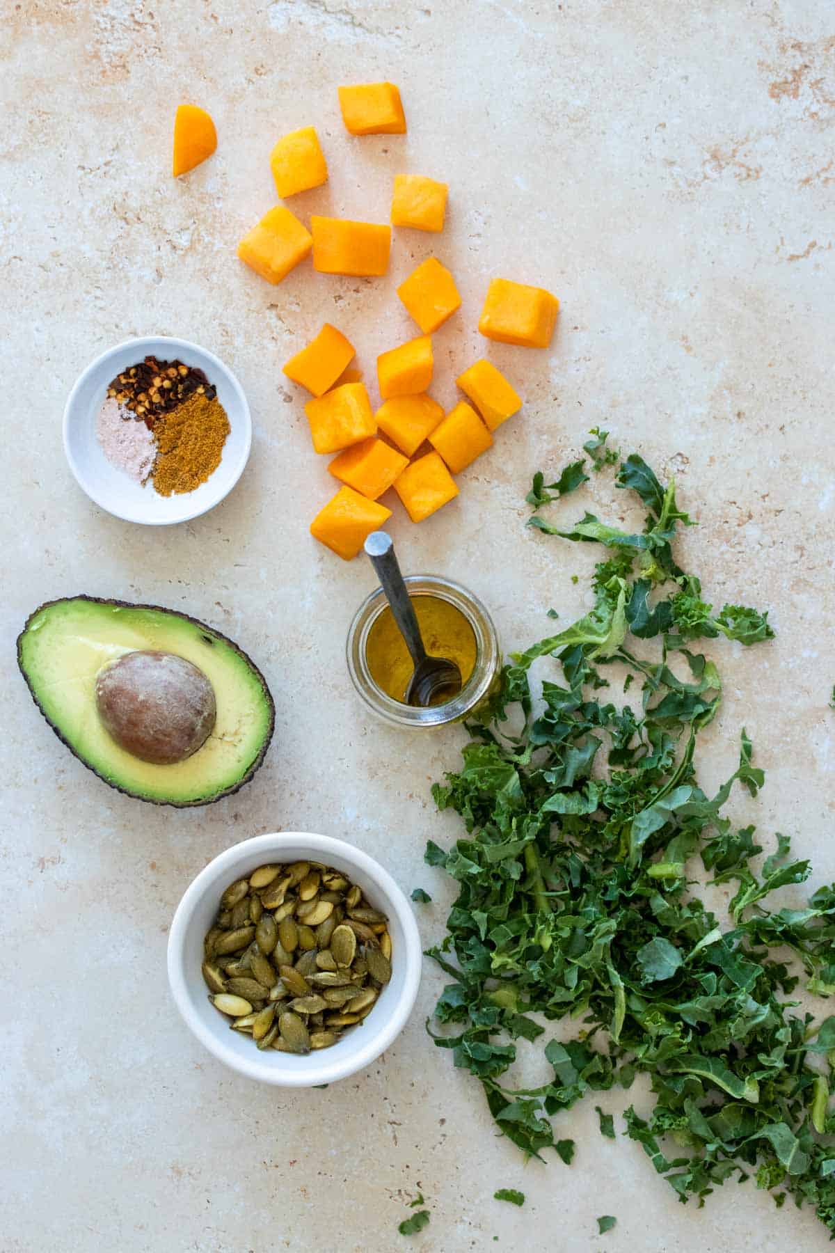 Tan surface with kale, butternut squash pieces, half an avocado, spices and olive oil on it