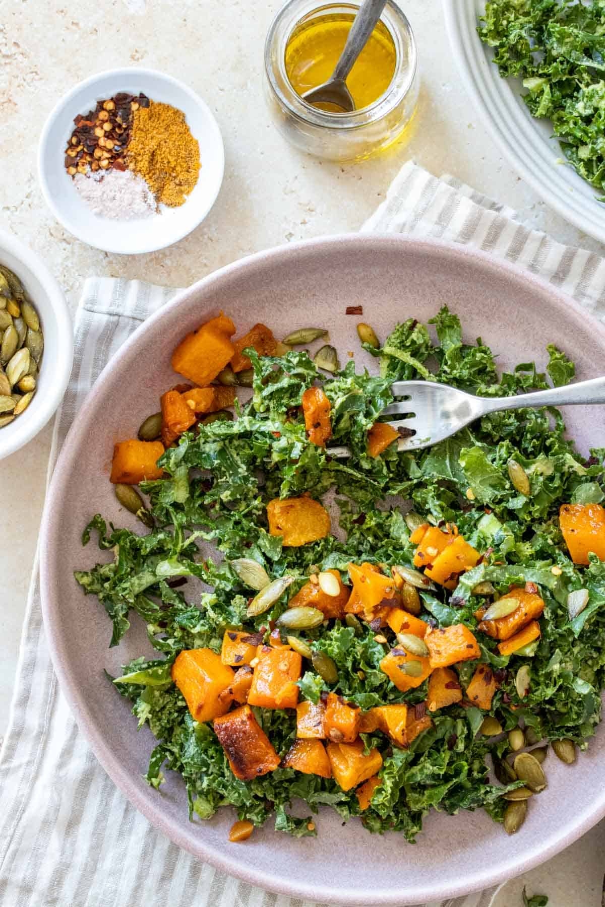 A fork getting a bite of a butternut squash kale salad on a pink plate sitting on a striped towel