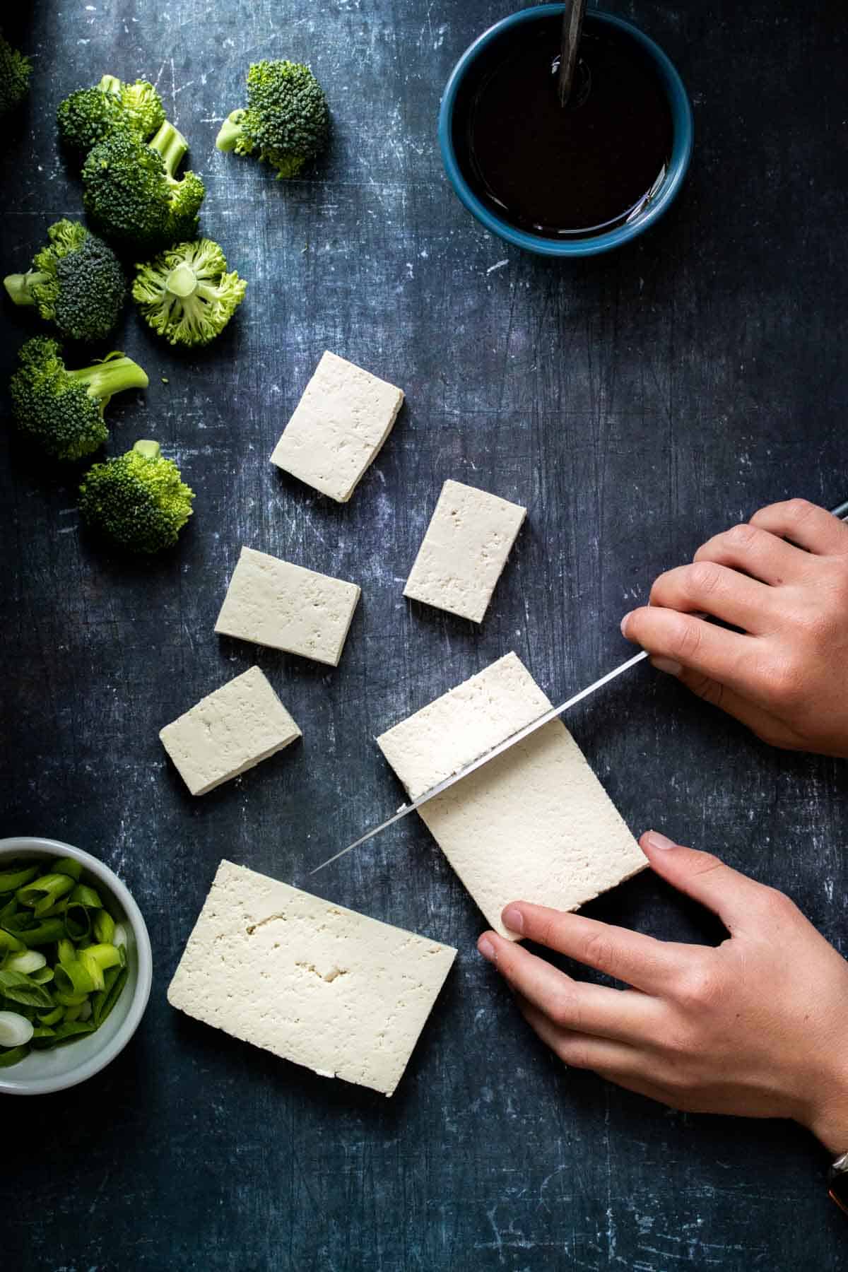 A hand holding a cutting a block of tofu into pieces on a dark surface next to veggies and sauce