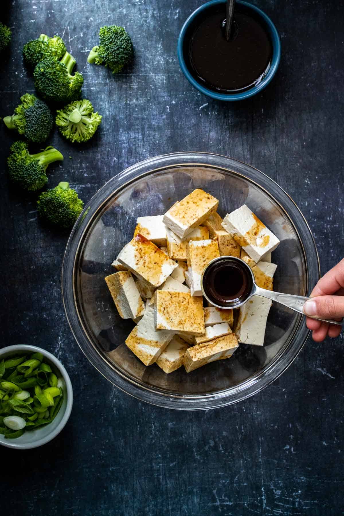 A hand putting teriyaki sauce from a tablespoon onto cubed tofu in a glass bowl