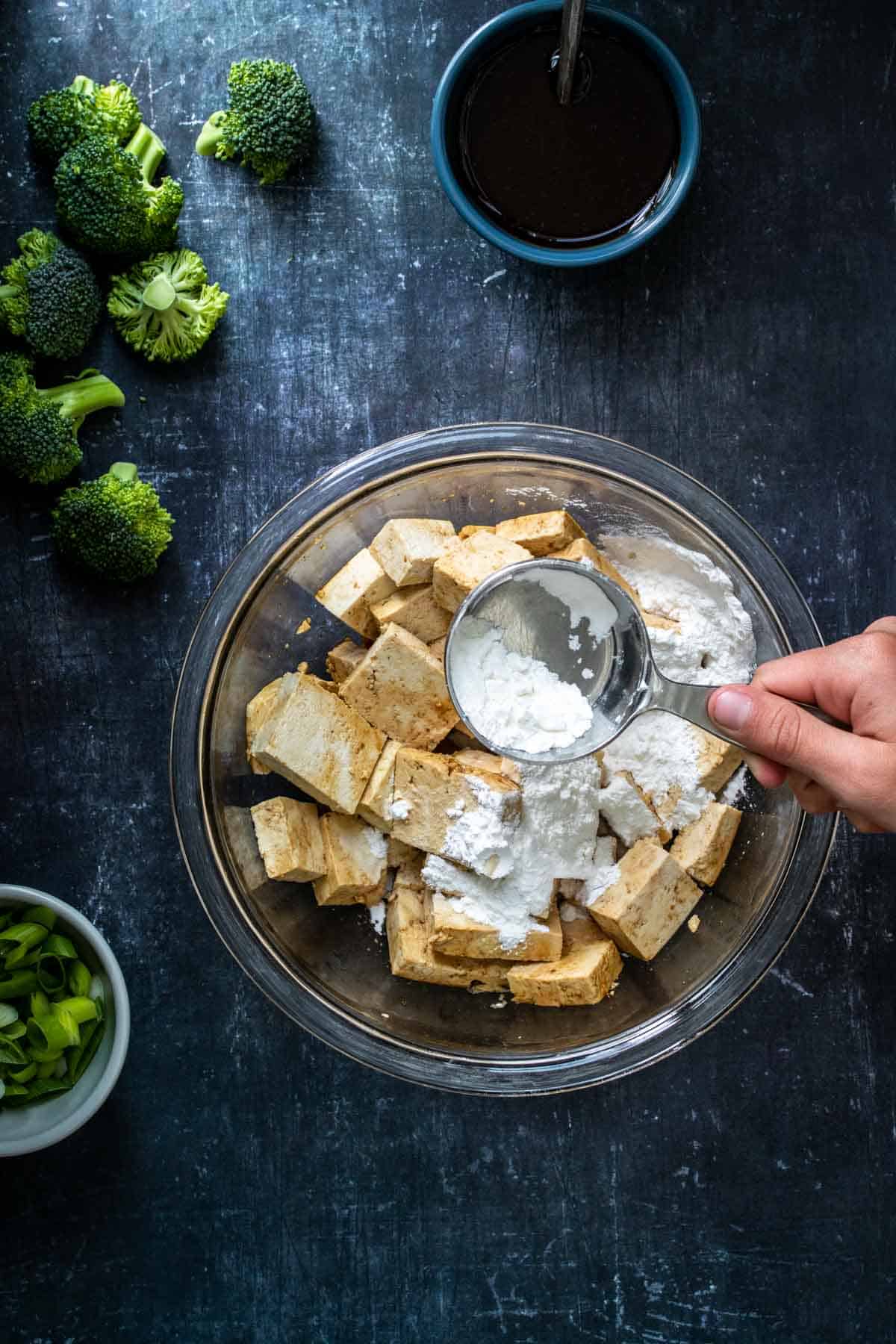 A hand pouring cornstarch from a metal measuring cup over cubed tofu covered in teriyaki sauce