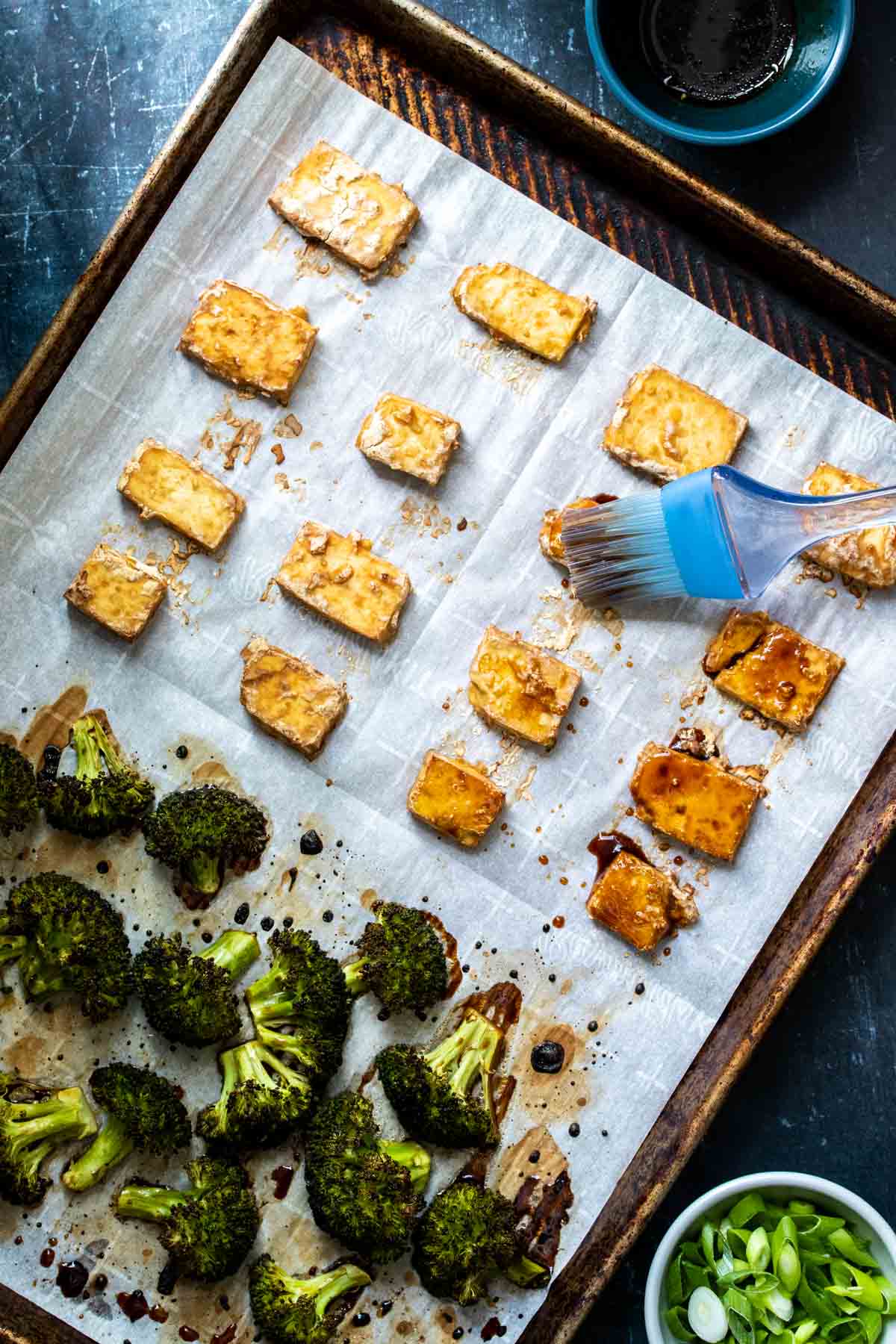 A pastry brush brushing teriyaki sauce over pieces of baked tofu on a parchment lined baking sheet with broccoli