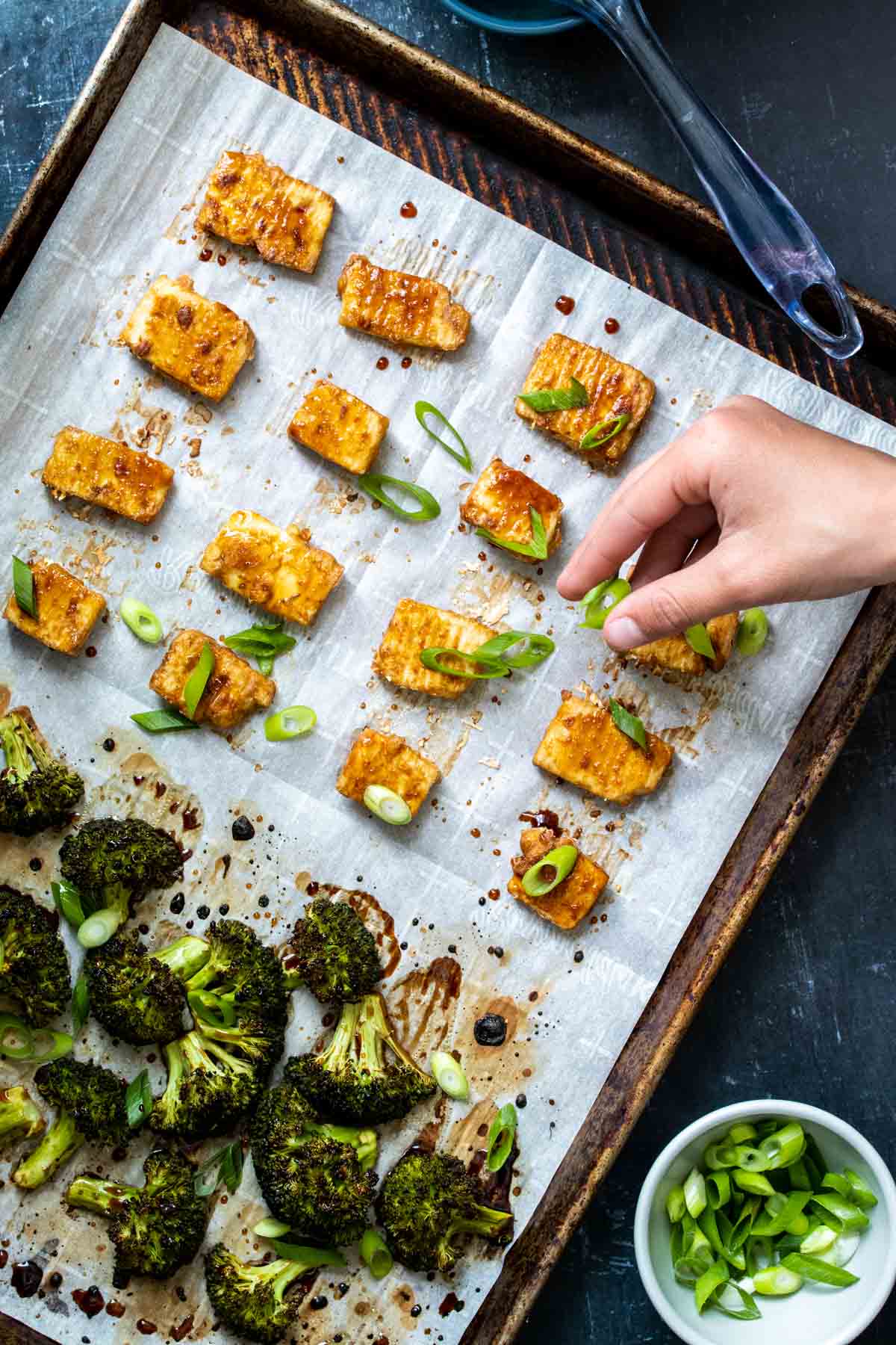 A hand sprinkling sliced green onions over tofu and broccoli pieces that are on a parchment lined baking sheet