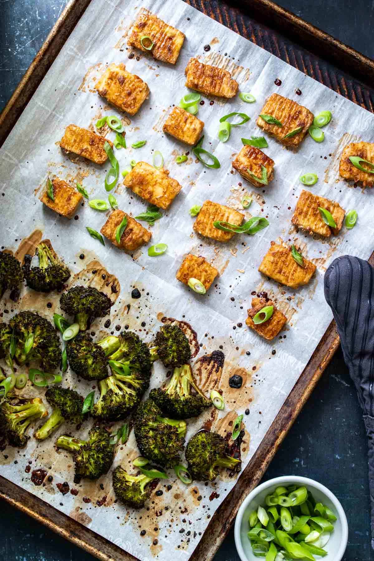 A baking sheet lined with parchment paper with baked tofu pieces and broccoli on it sprinkled with green onions