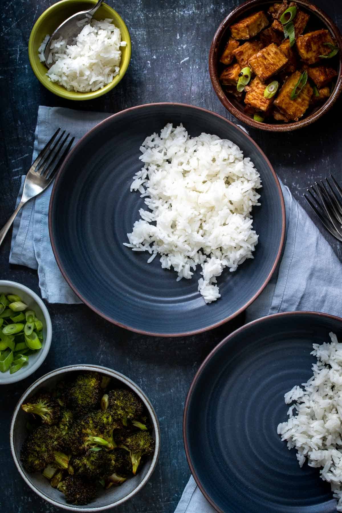 A dark blue bowl with rice in it surrounded by ingredients for tofu teriyaki