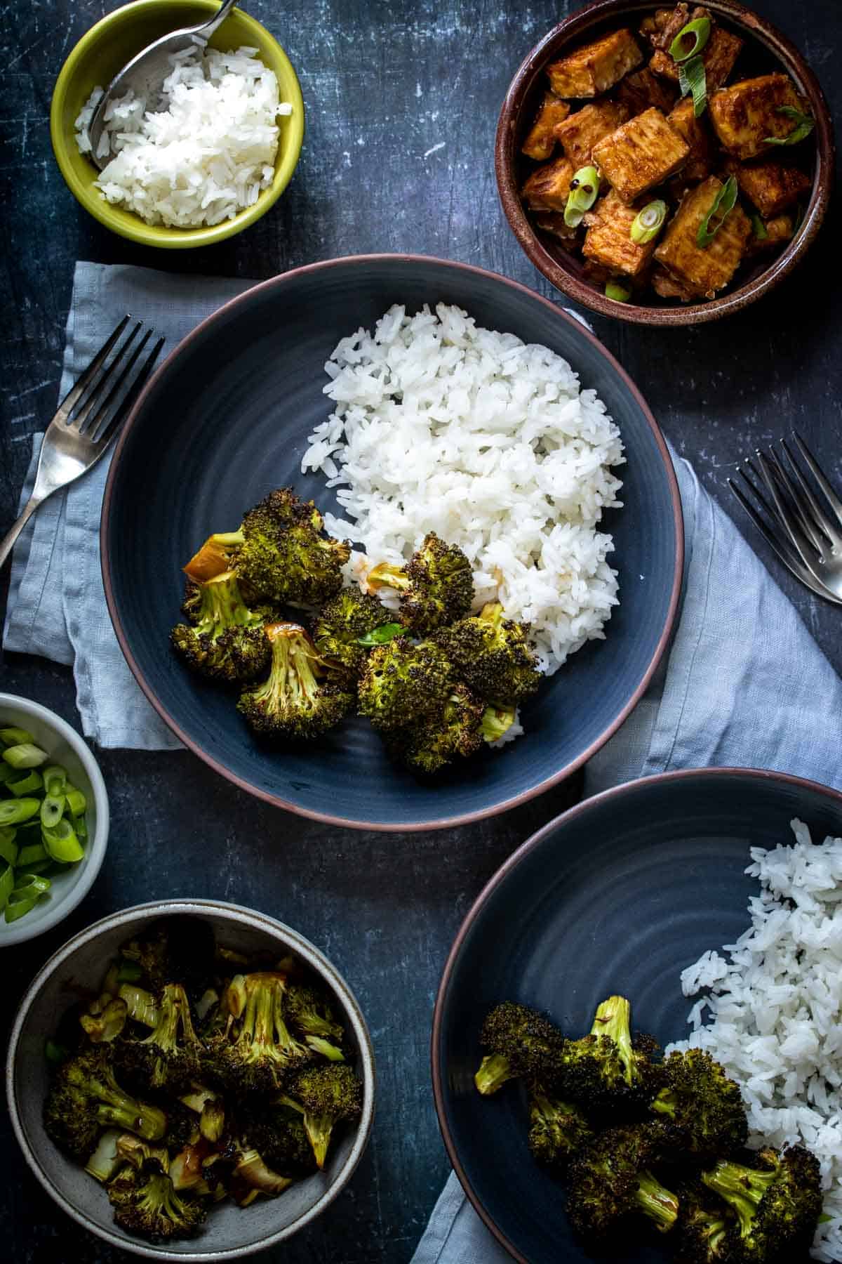 Broccoli and white rice in a dark blue bowl surrounded by ingredients to make tofu teriyaki bowls