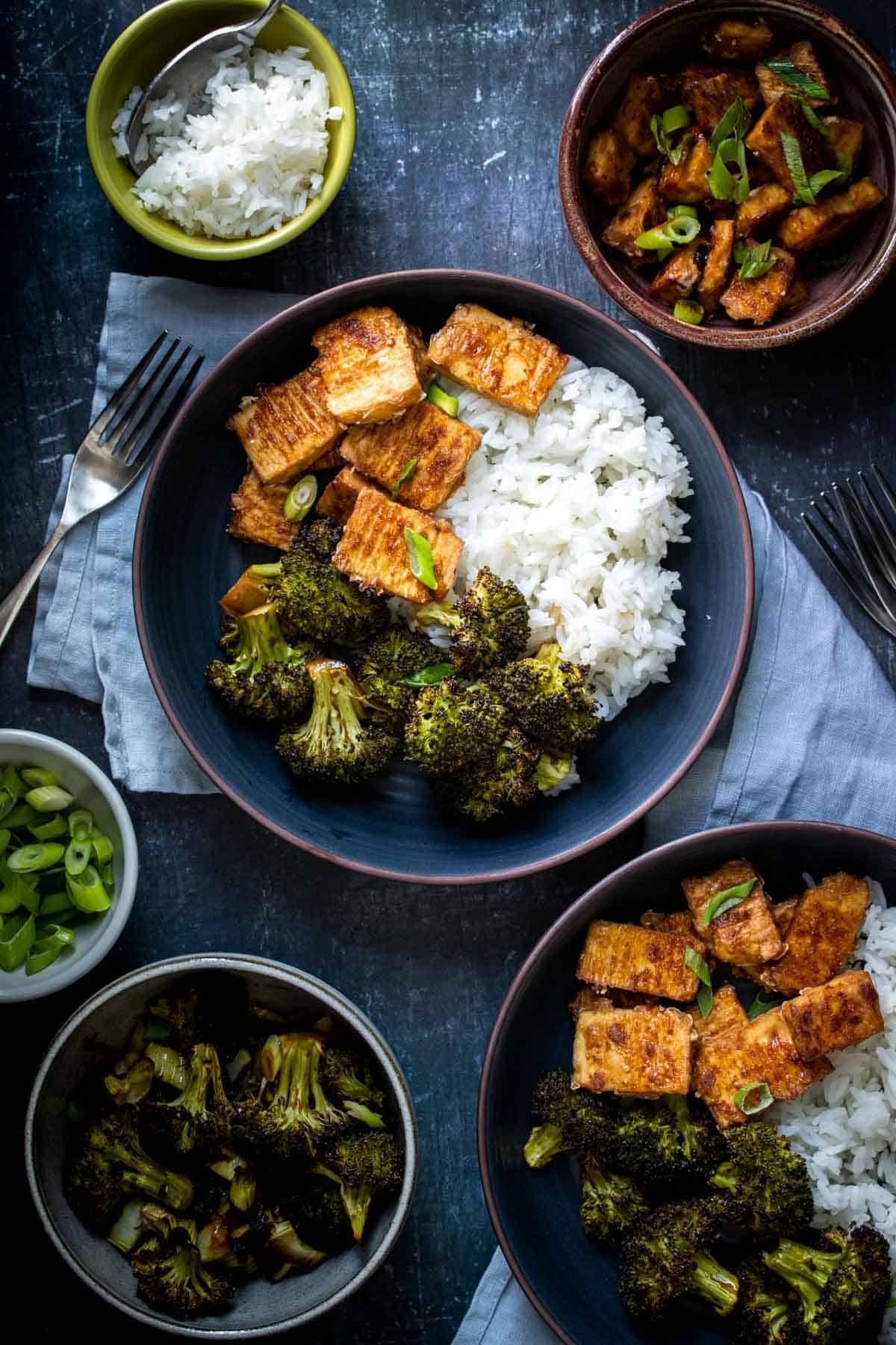 Teriyaki covered tofu pieces in a dark blue bowl next to rice and broccoli