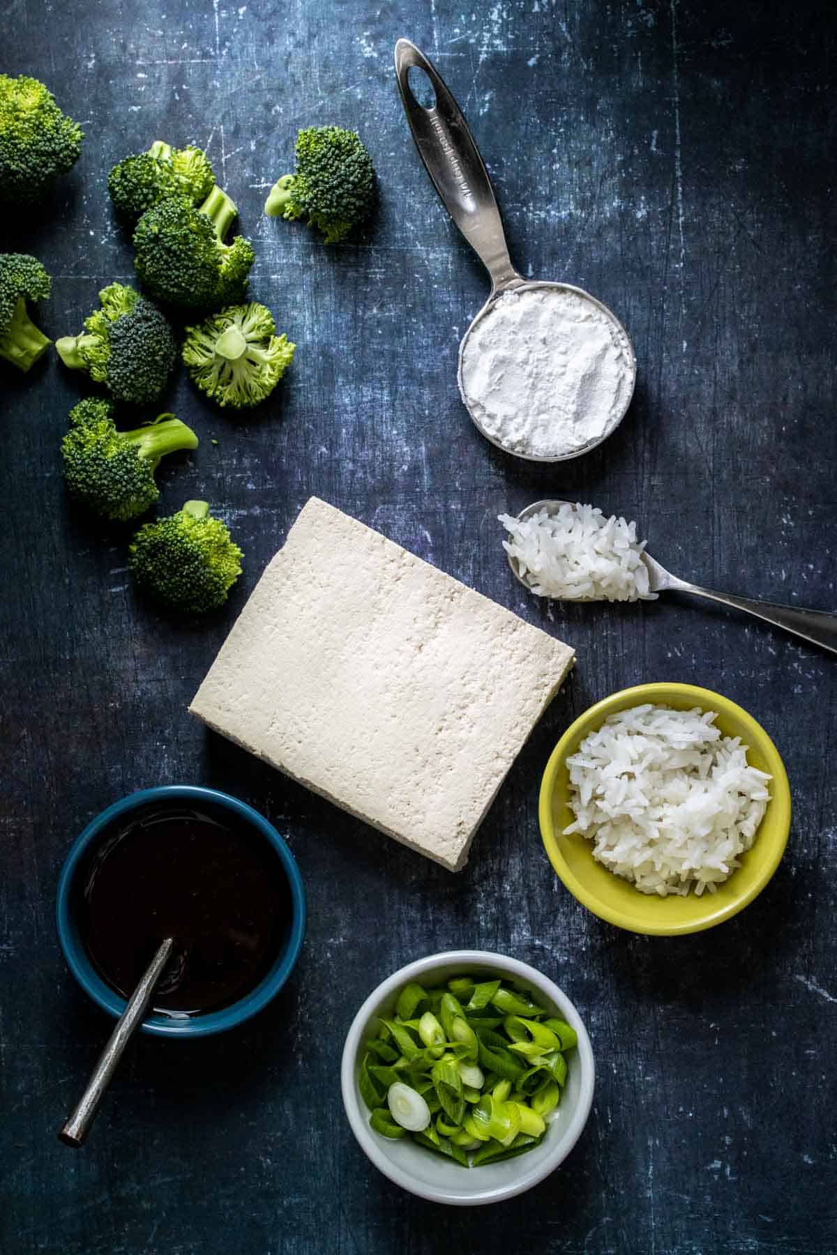 A block of tofu, broccoli, green onions, rice, teriyaki sauce and cornstarch on a dark grey surface