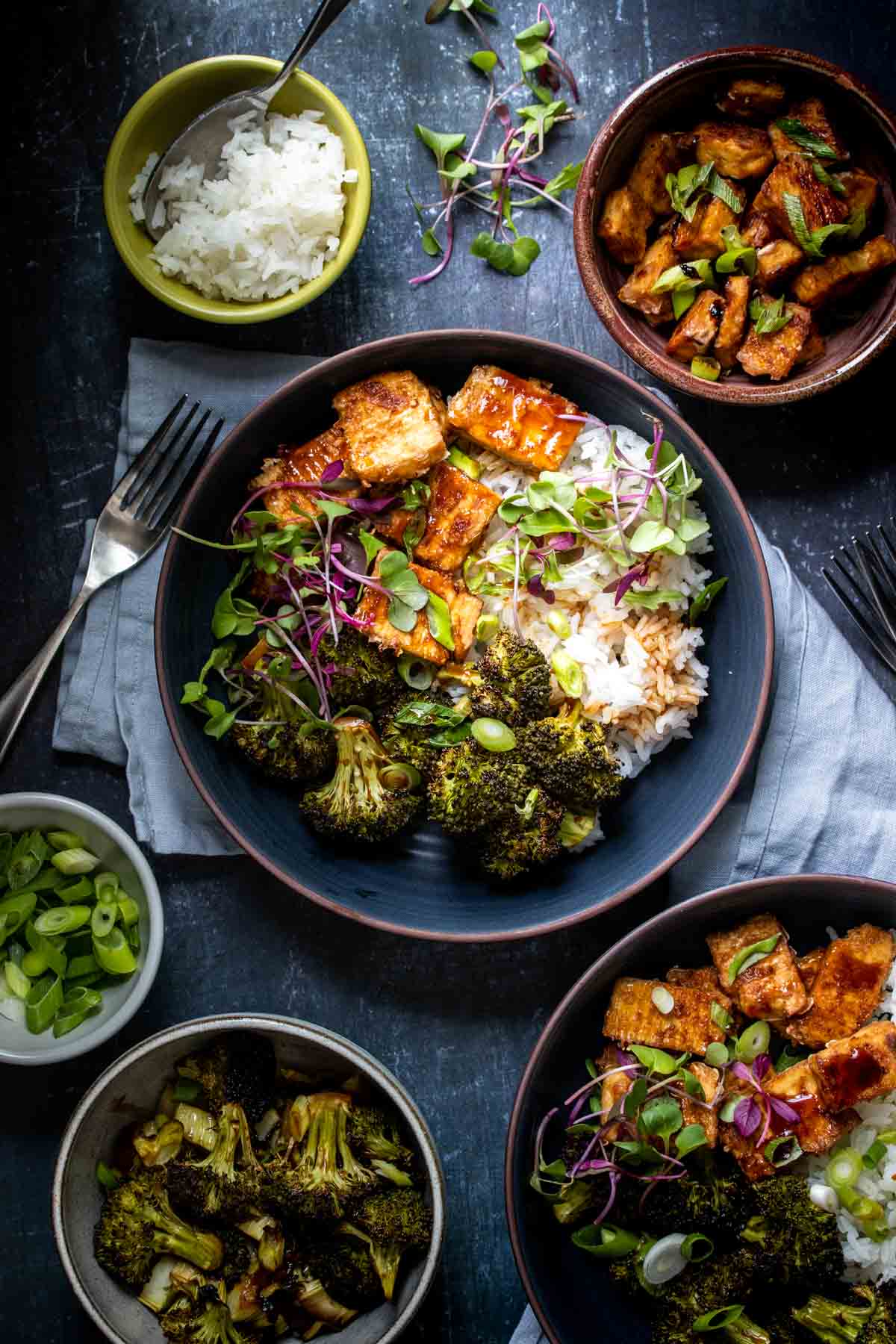 A dark blue bowl filled with baked teriyaki tofu and broccoli over white rice surrounded by more ingredients