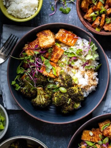 Teriyaki tofu and broccoli sprinkled with green onions and microgreens over rice in a dark blue bowl