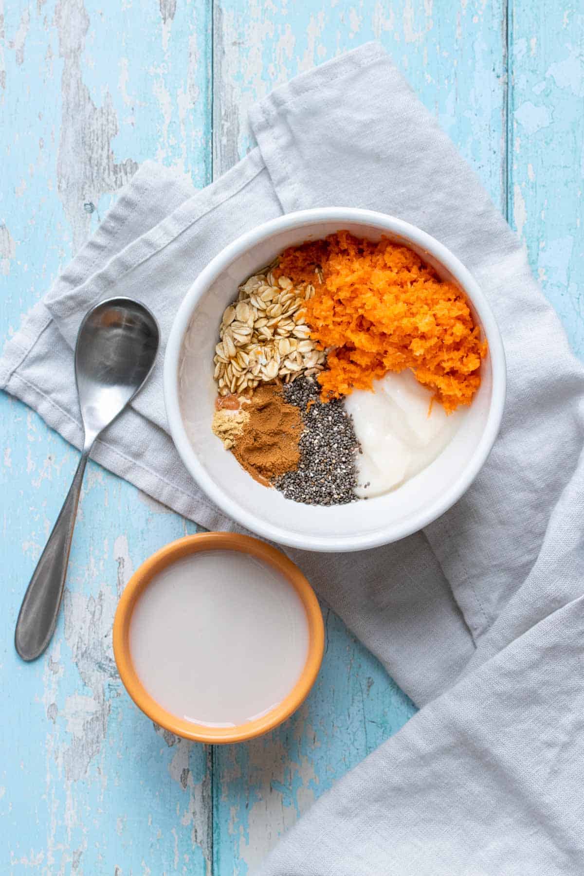A large white bowl with oats, carrots, spices and yogurt inside next to a smaller bowl with milk
