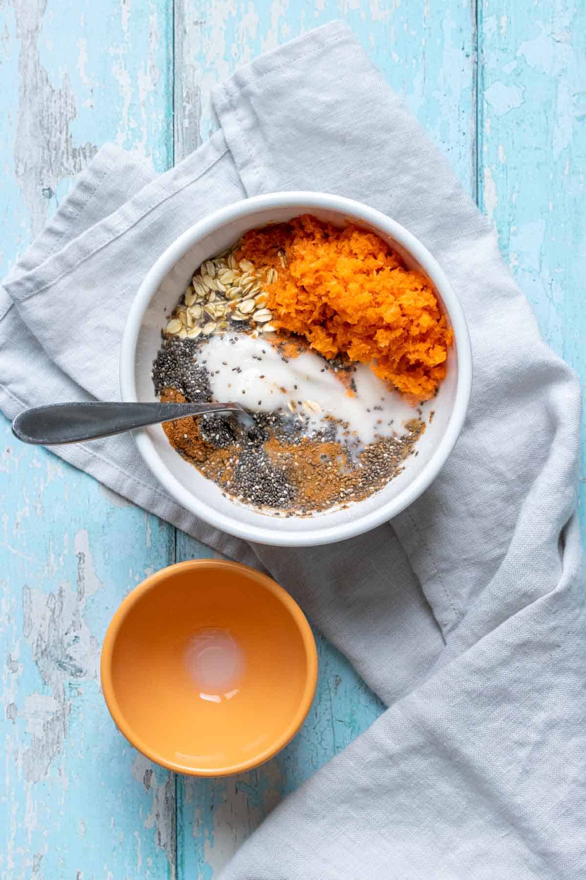 A large white bowl with ingredients for carrot cake overnight oats and a spoon next to a smaller orange bowl
