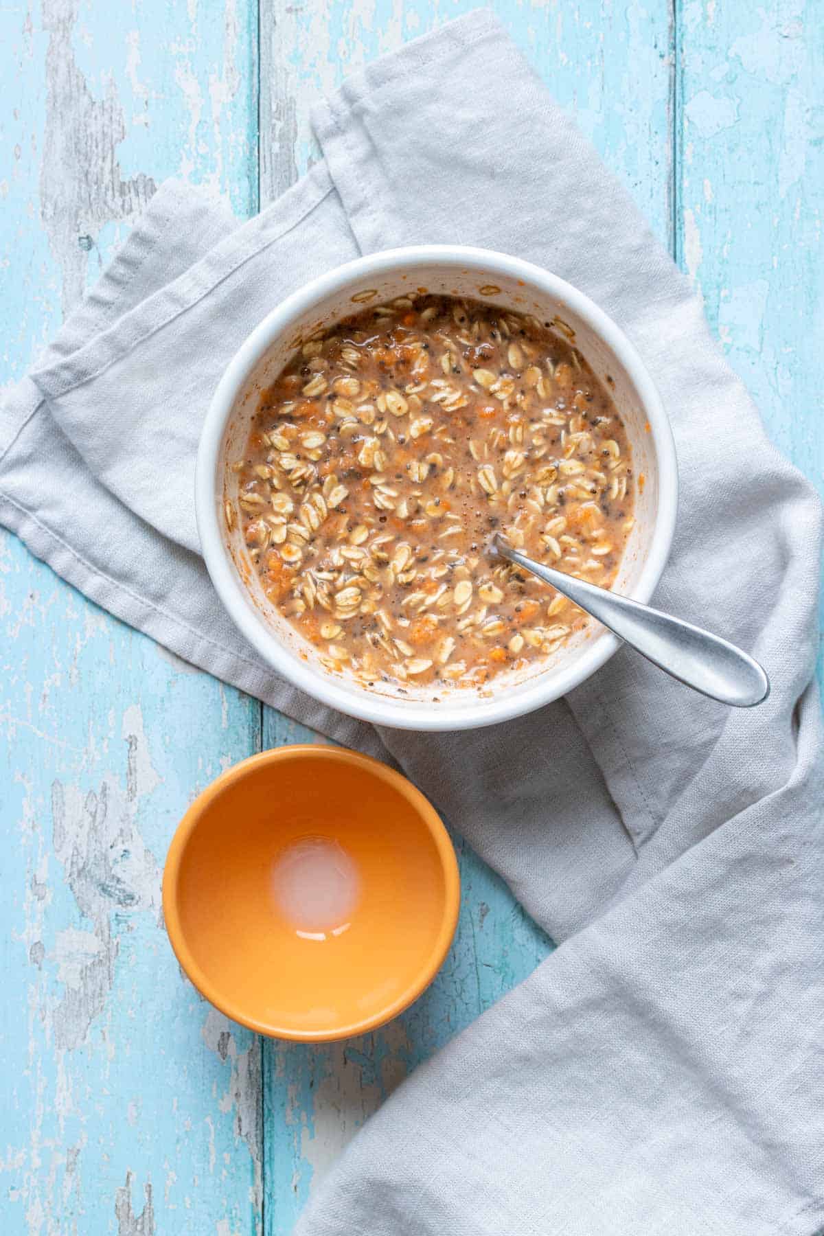 A large white bowl with oats mixed with milk and carrots next to a small orange bowl on a blue surface