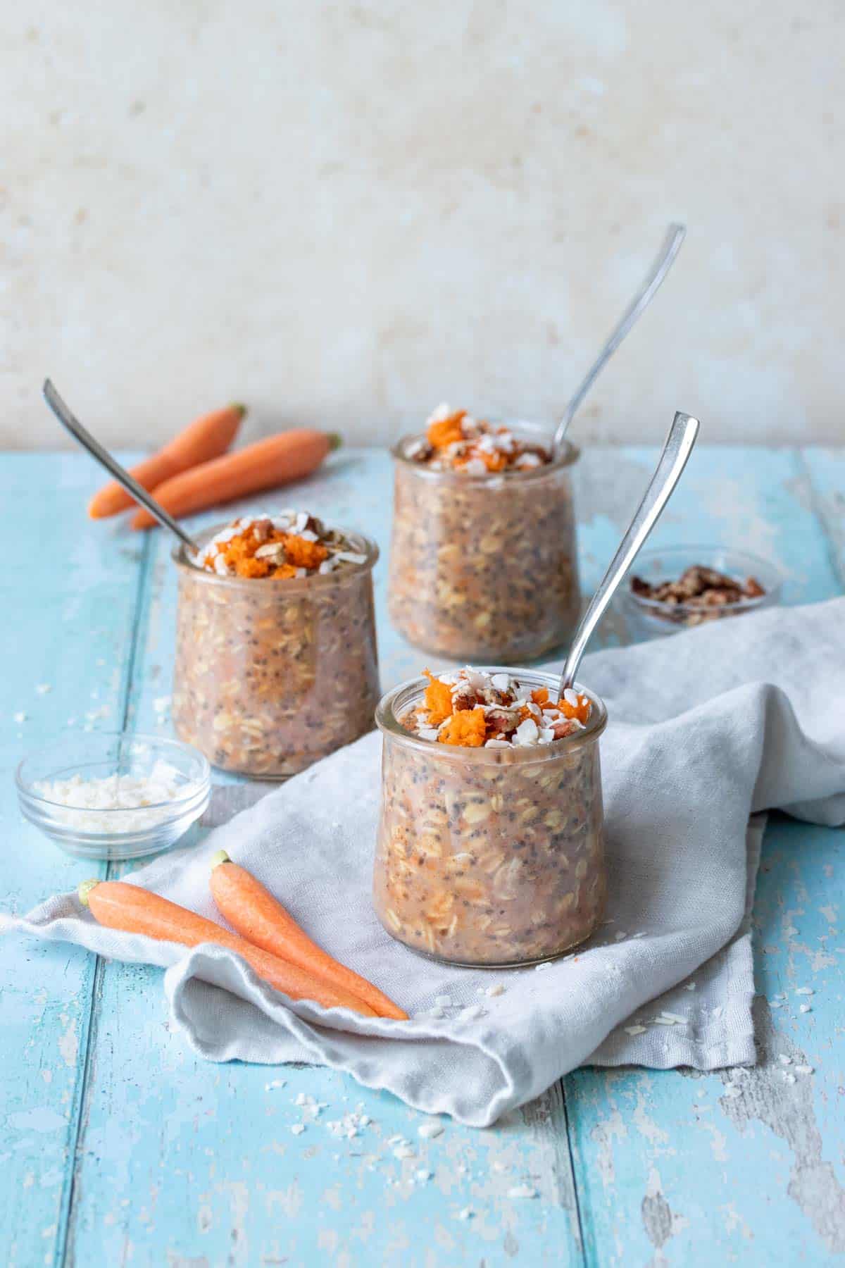 Three jars with carrot cake overnight oats on a grey towel on a blue wooden surface