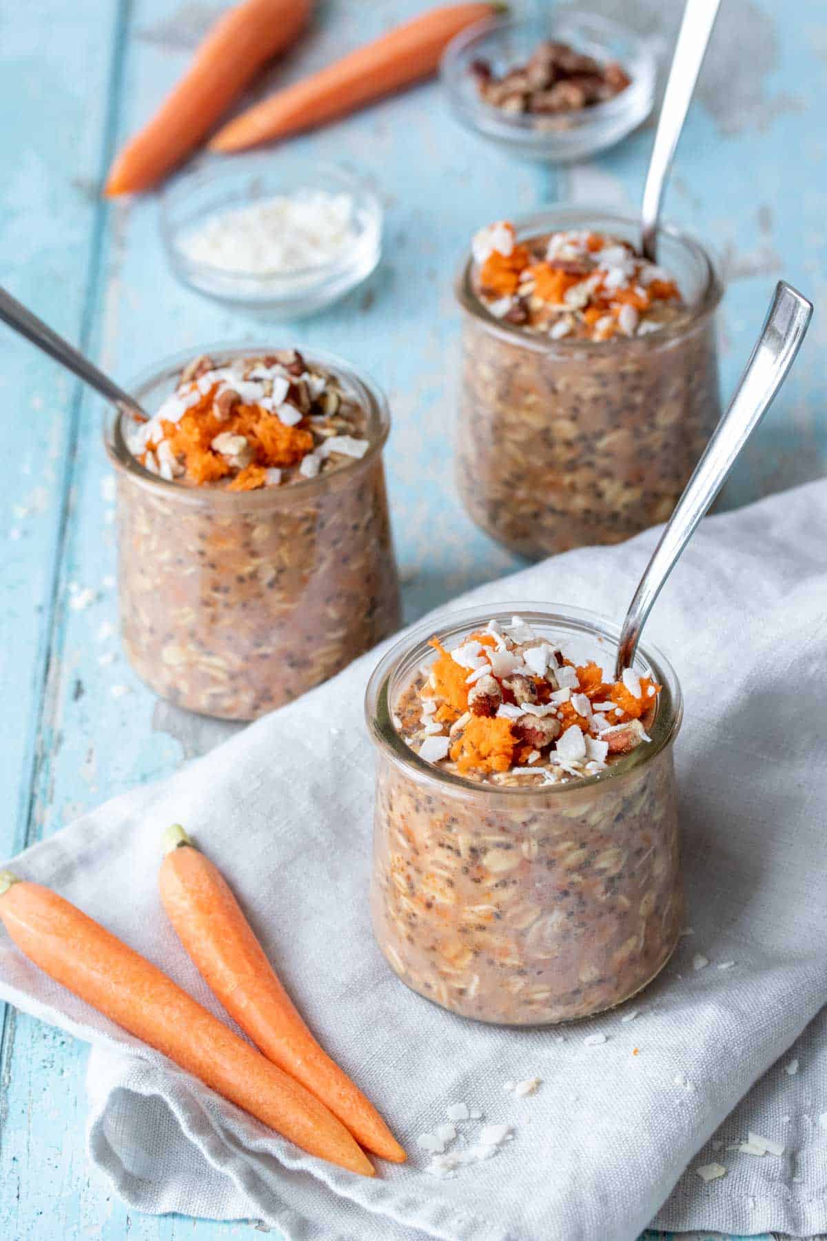 Three jars with a carrot oat mixture and spoons sitting on a grey towel on a blue wooden surface next to whole carrots