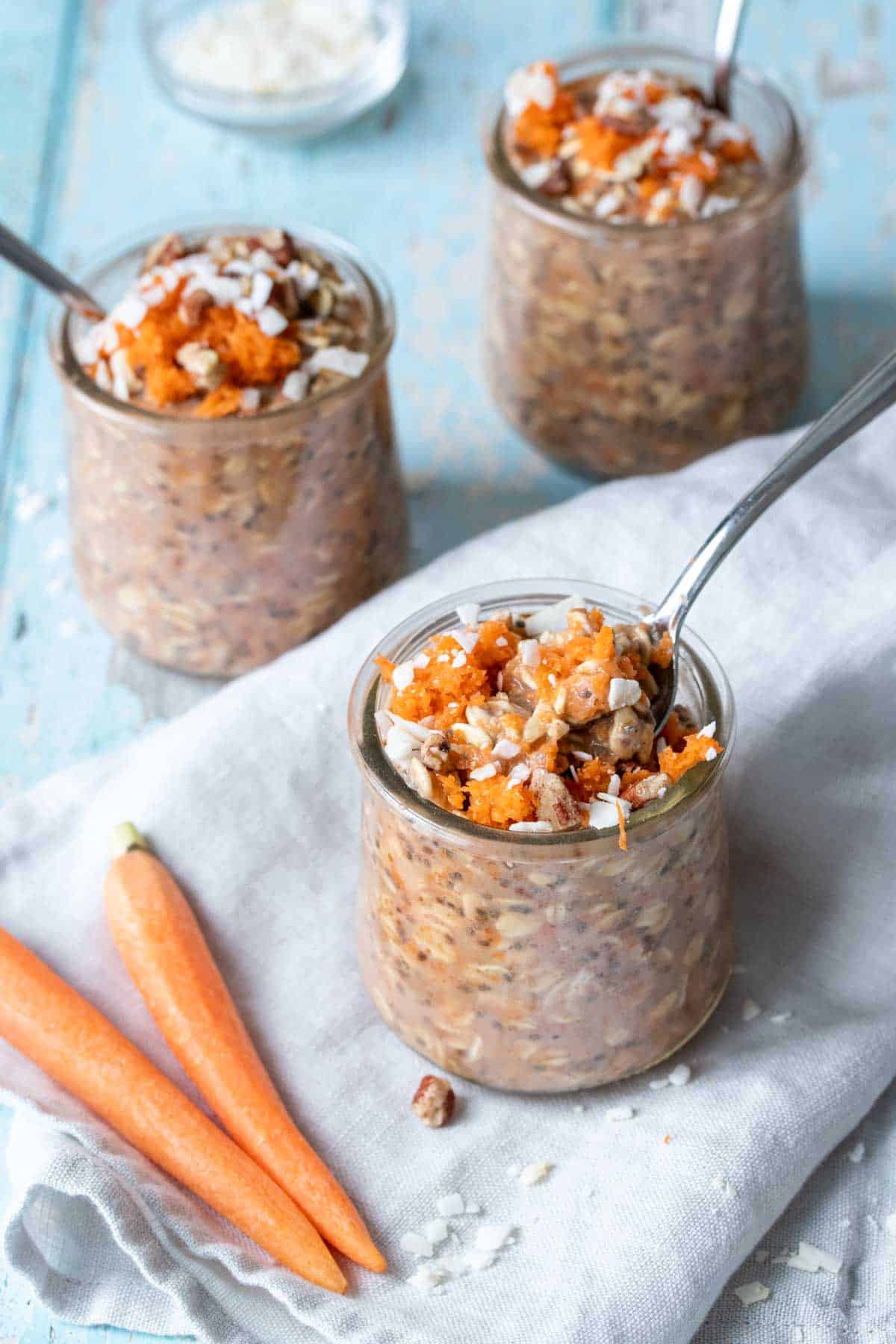 A spoon coming out of a glass jar sitting on a grey towel with a scoop of carrot cake oats