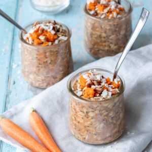 A blue surface with a grey towel and glass jars on it filled with carrot cake oats and spoons inside