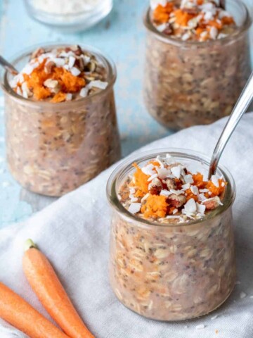 A blue surface with a grey towel and glass jars on it filled with carrot cake oats and spoons inside.