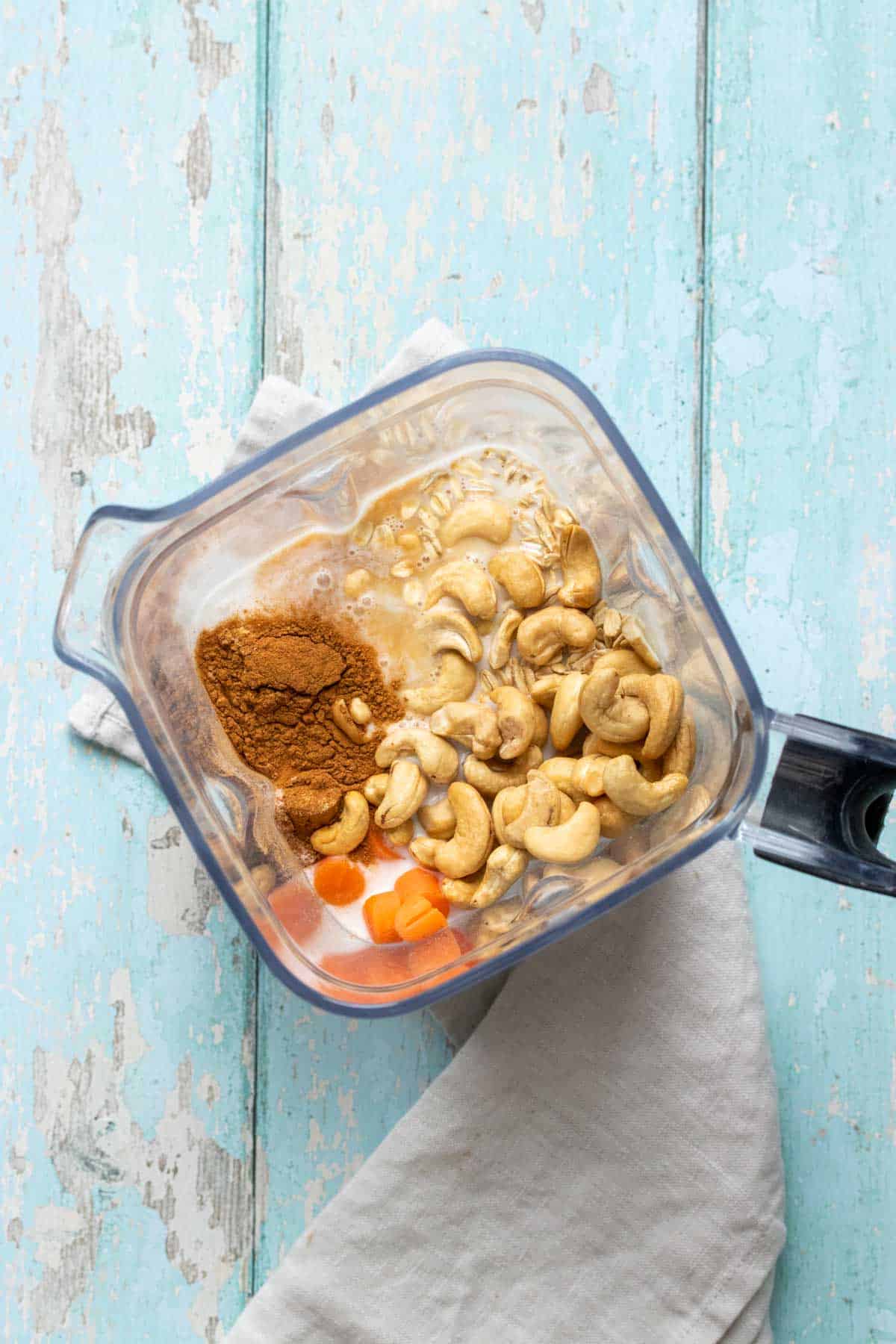 Top view of a blender on a blue wooden surface with cashews, carrots, spices and milk inside