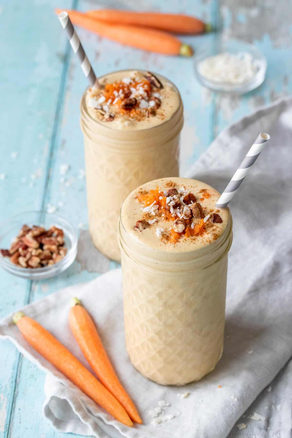 Two tall glass jars with peach colored smoothies topped with carrots, pecans and coconut