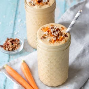 A tall glass jar in front of another jar both filled with peach colored smoothies topped with shredded carrots, chopped pecans and coconut.