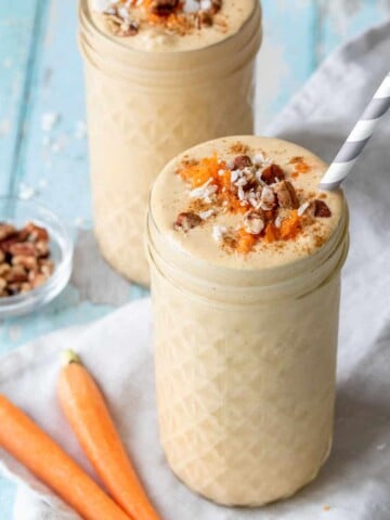 A tall glass jar in front of another jar both filled with peach colored smoothies topped with shredded carrots, chopped pecans and coconut.