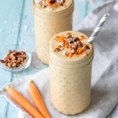 A tall glass jar in front of another jar both filled with peach colored smoothies topped with shredded carrots, chopped pecans and coconut